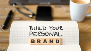 Closeup on notebook over vintage desk surface, front focus on wooden blocks with letters making Build Your Personal Brand text. Business concept image with office tools and coffee cup in background