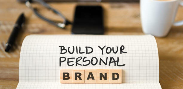 Closeup on notebook over vintage desk surface, front focus on wooden blocks with letters making Build Your Personal Brand text. Business concept image with office tools and coffee cup in background