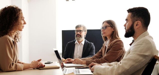 Four professionals seated around a conference table with laptops and notebooks, engaged in a meeting.
